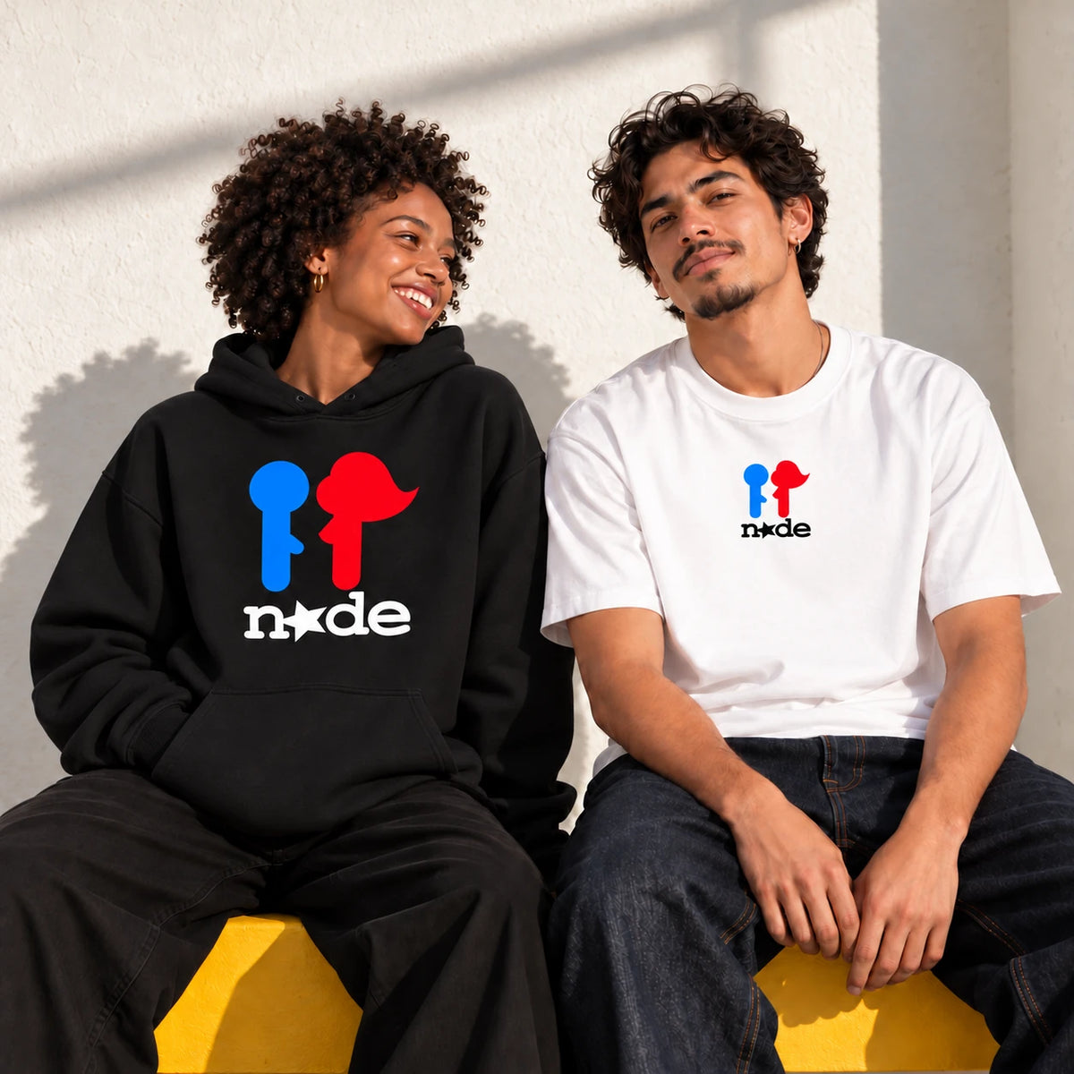 Two adult models wearing the official NXDE logo on a black hoodie and white tee, seated on a yellow bench
